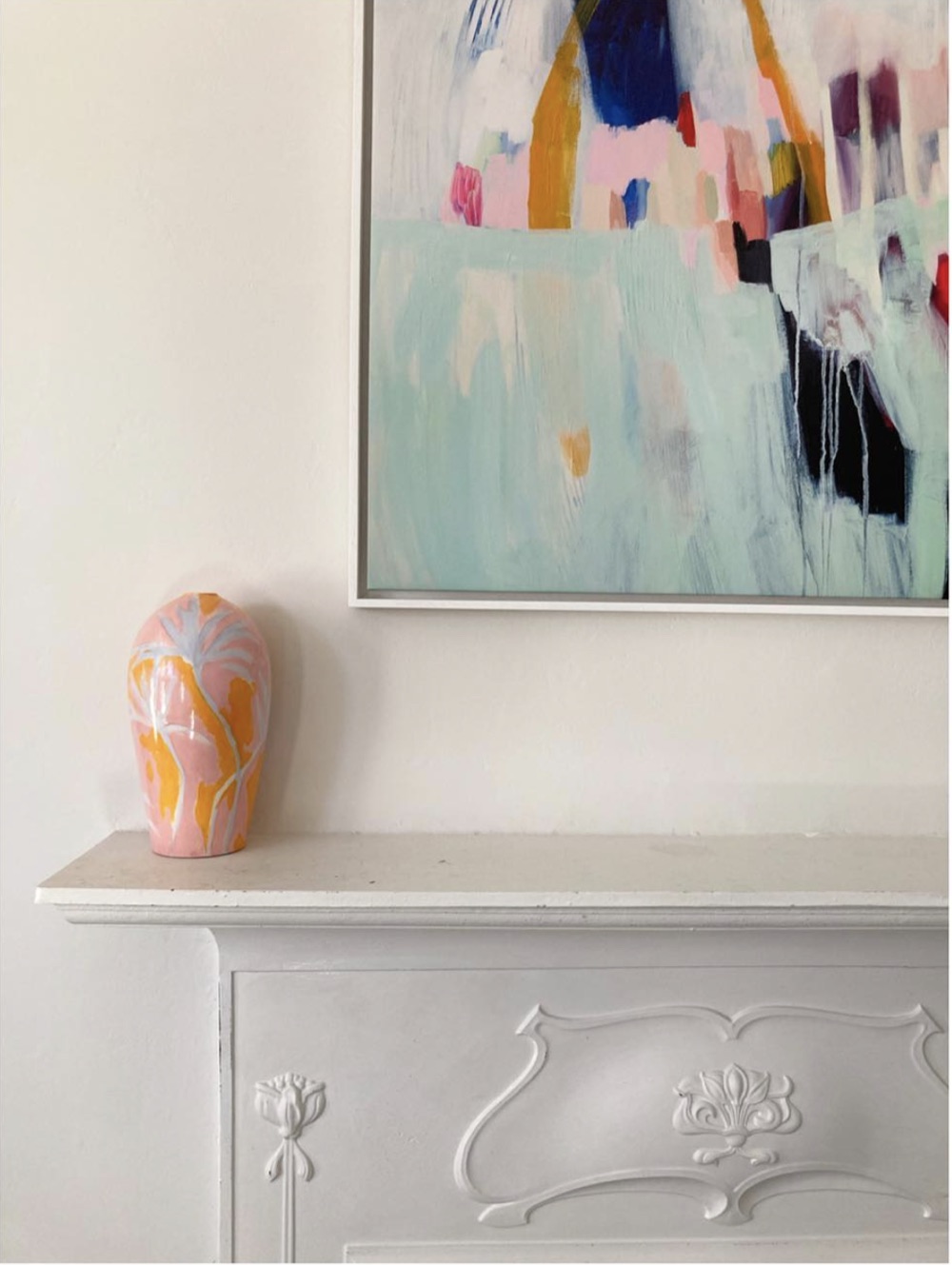 A pink and orange ceramic vase on a white mantelpiece beneath an abstract painting